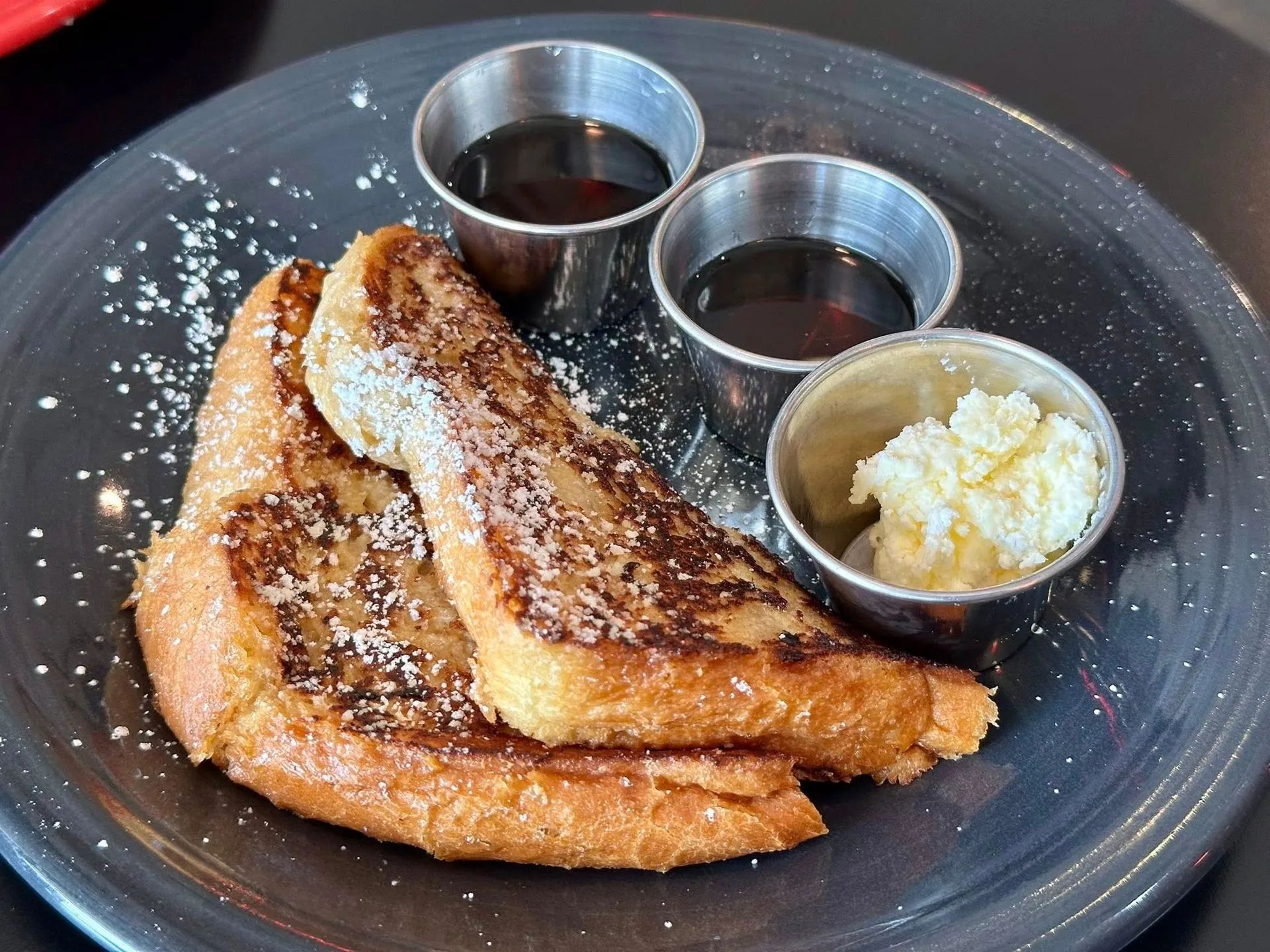 Two slices of golden-brown French toast dusted with powdered sugar, served with side cups of syrup and butter.