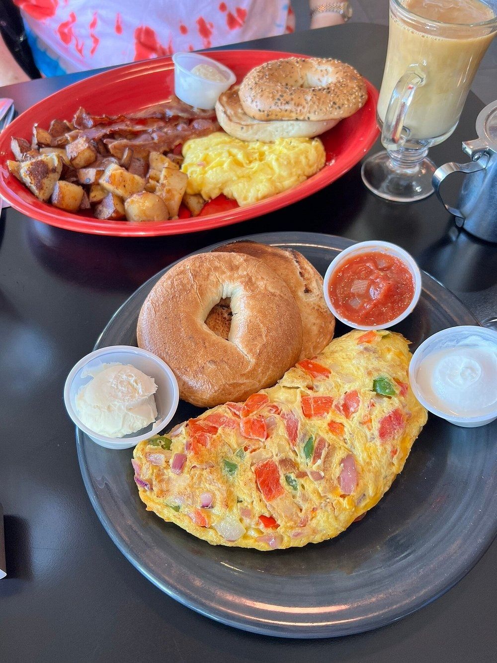 Two breakfast plates with omelets, bagels, potatoes, and drinks on a café table