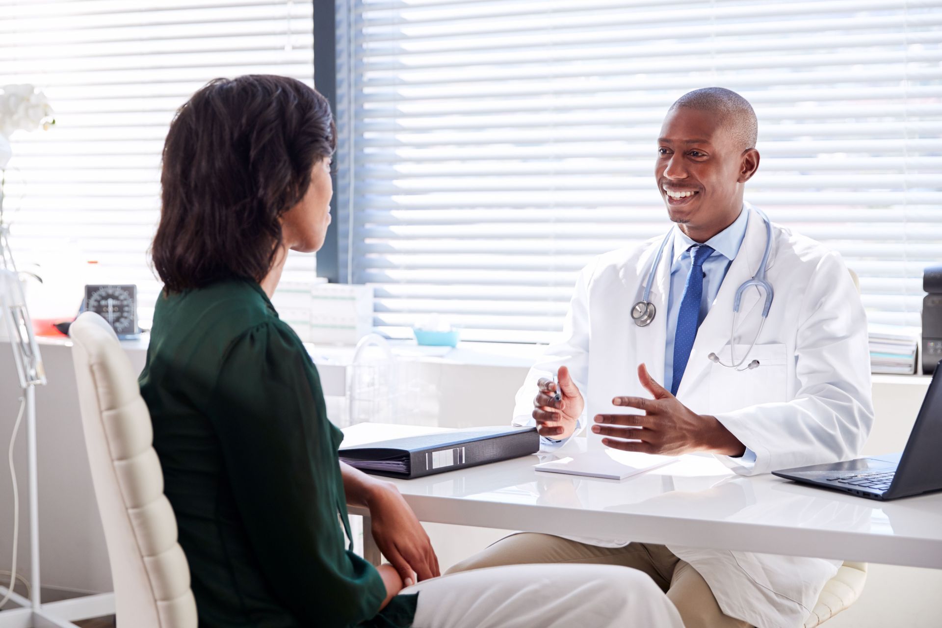 Doctor in white coat smiles, talking with patient at desk in office setting.