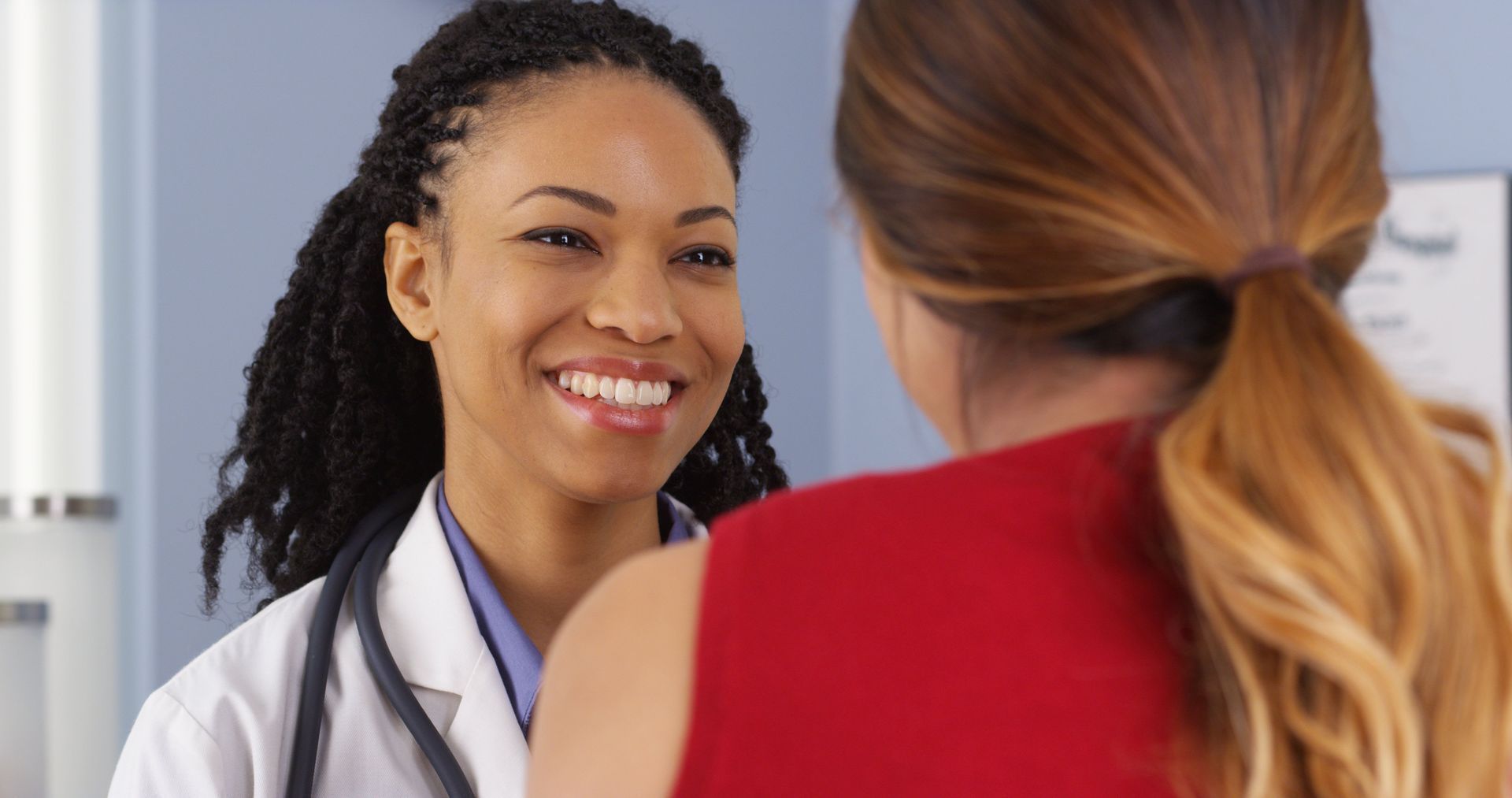 Doctor smiling at a patient in an office. Doctor wears a white coat and stethoscope. Patient wears red.