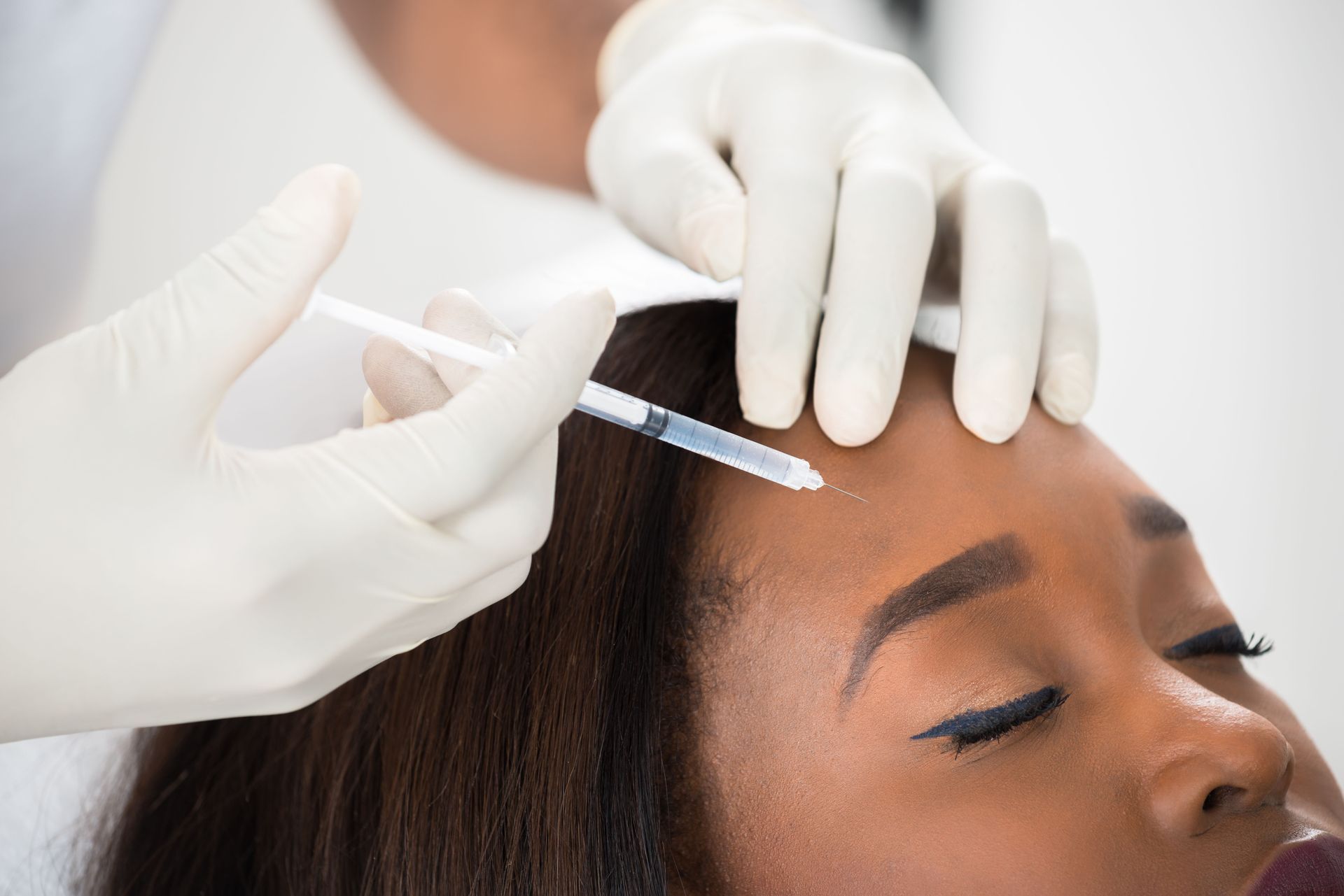 Person receiving a cosmetic injection in the forehead; white-gloved hands hold a syringe.
