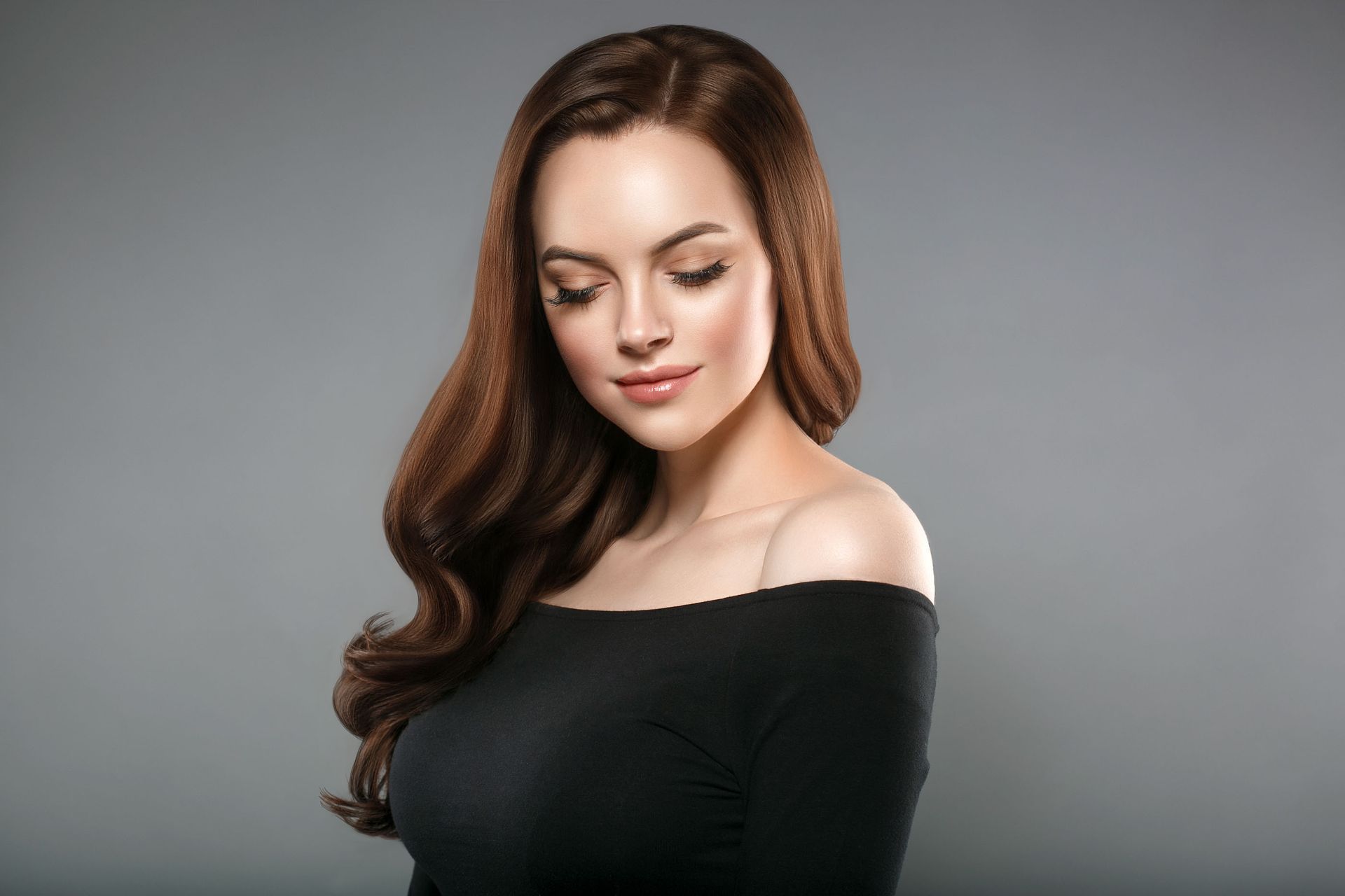 Woman with wavy brown hair, wearing a black off-the-shoulder top, looking down. Gray background.