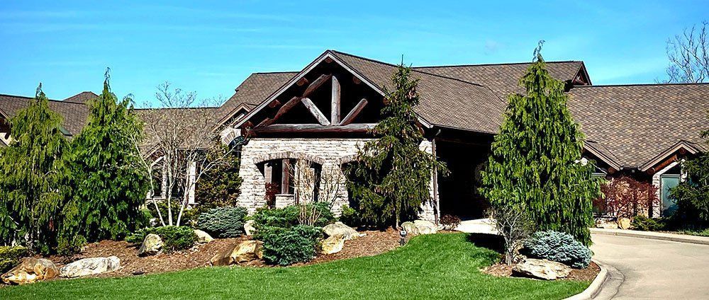 Stone house with brown roof, green lawn, trees, and blue sky.