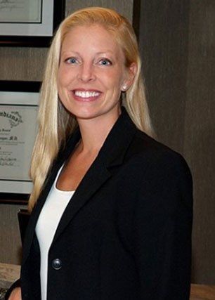 Woman with long blonde hair smiles, wearing a black blazer and white top, in an office setting.