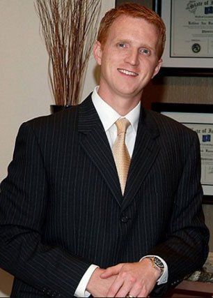 Man in suit smiling, leaning on a desk, with diplomas in the background.
