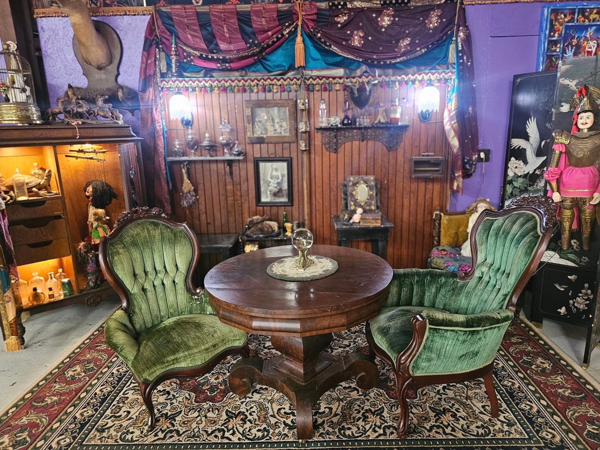 Vintage living room with green armchairs, a round wooden table, ornate decor, and purple curtains.