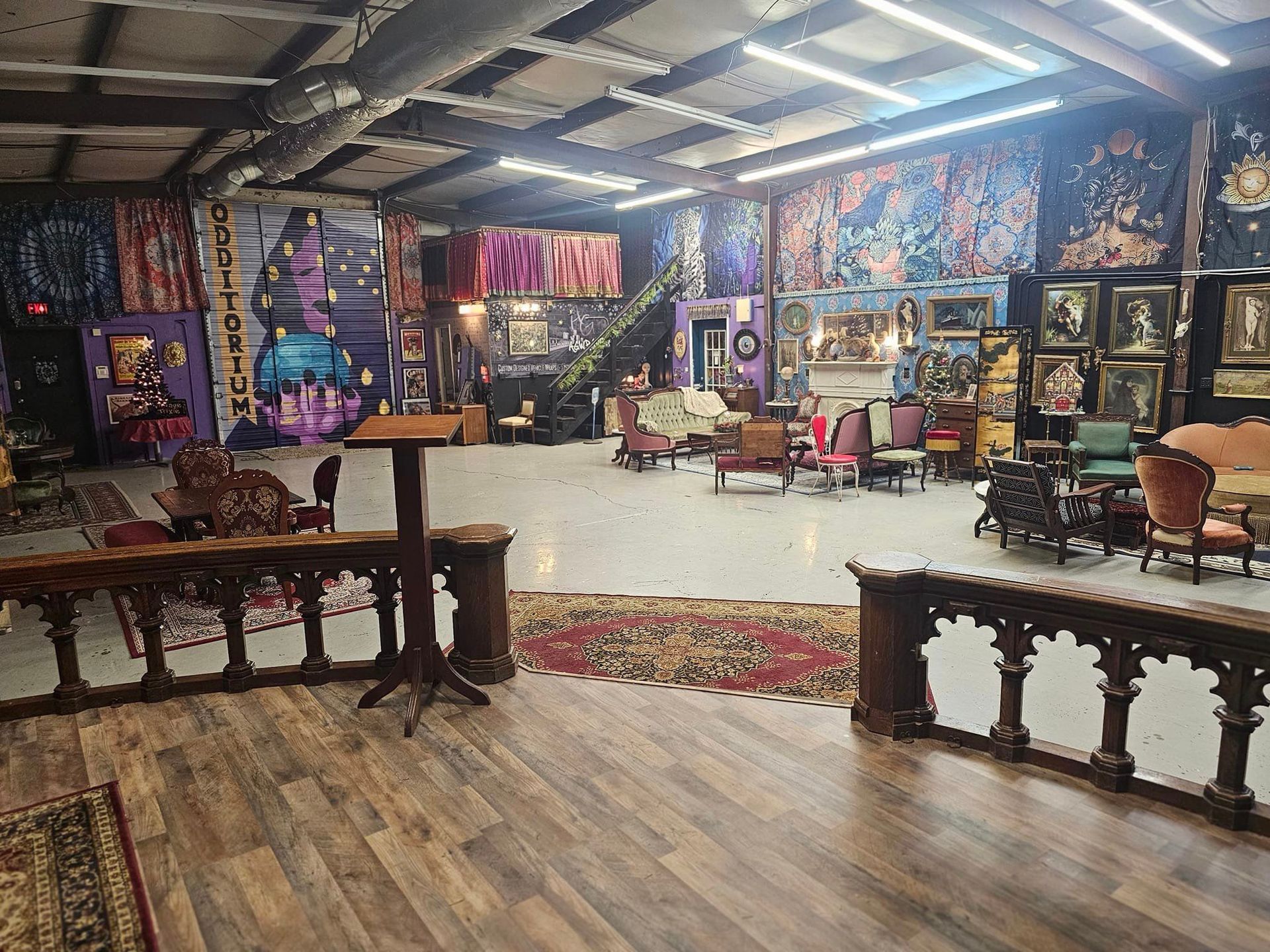 Colorful art-filled room with wooden tables, patterned rugs, and chairs under a high ceiling