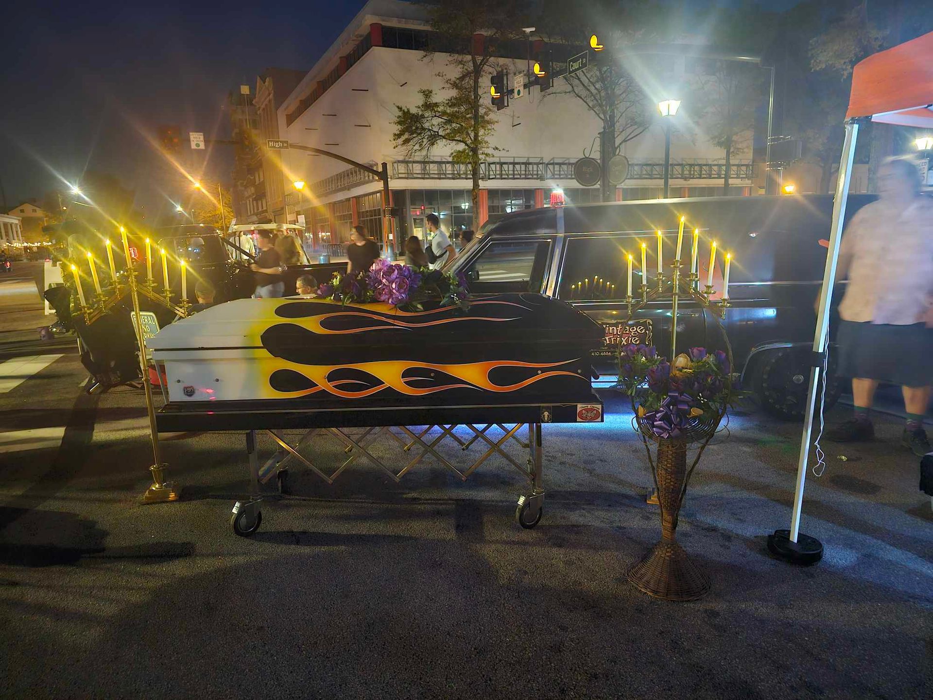 Decorated casket with flame design at a nighttime outdoor memorial, lit by candles and flowers