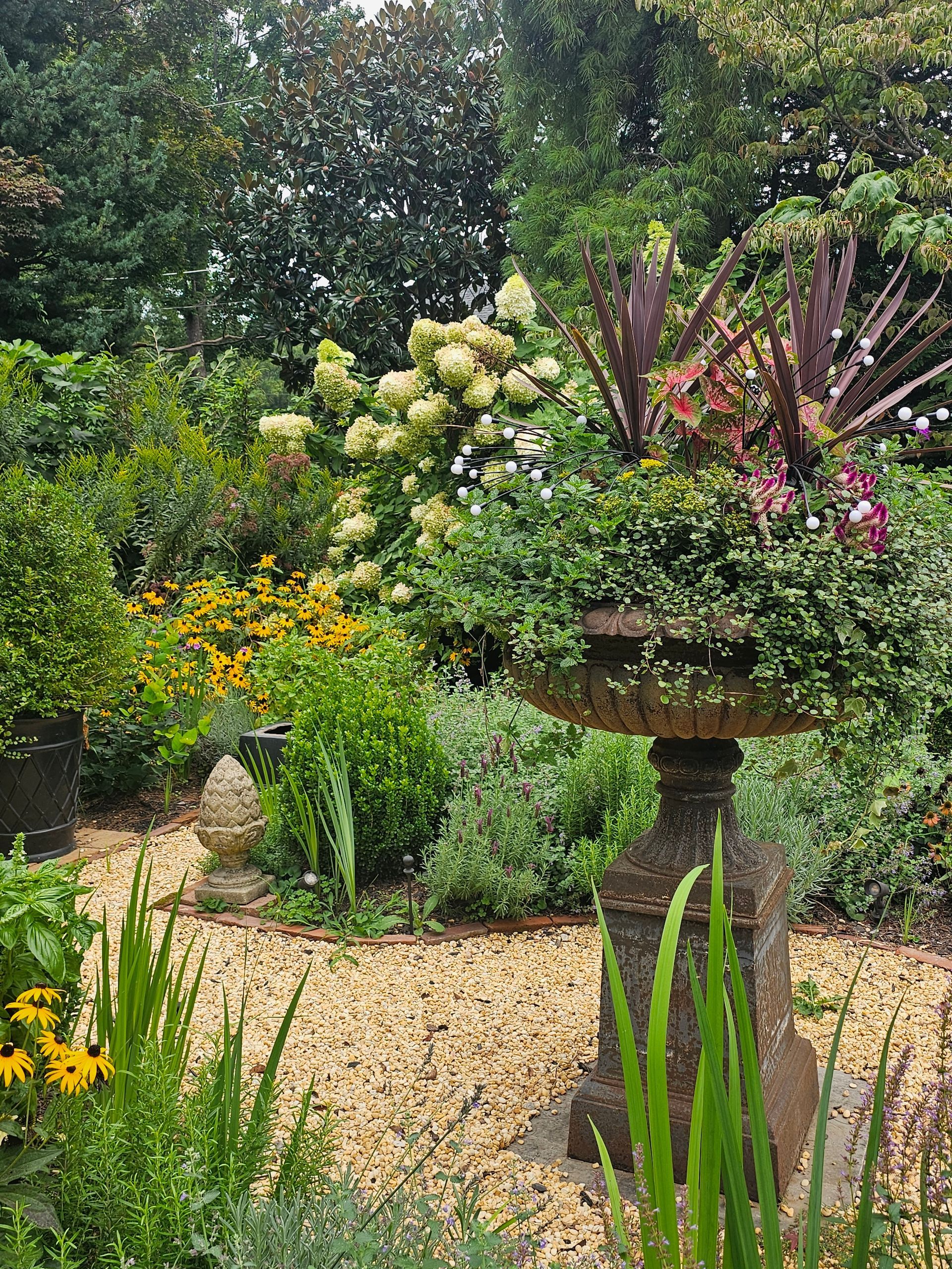 Lush garden with flowering shrubs, green foliage, and a stone birdbath beside a gravel path.