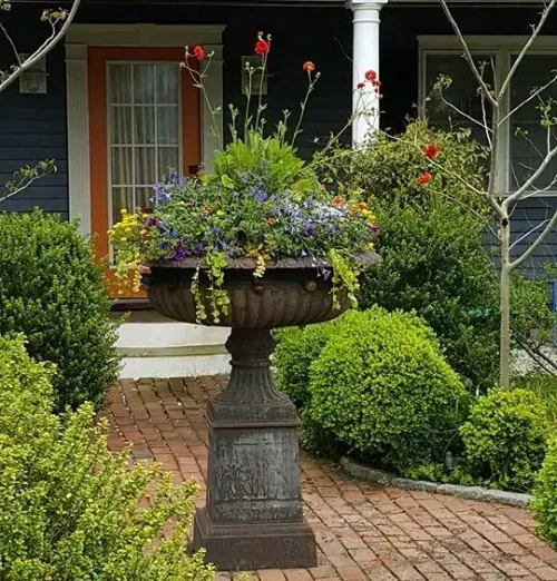 Flower-filled urn planter on a brick path in a garden beside a house porch