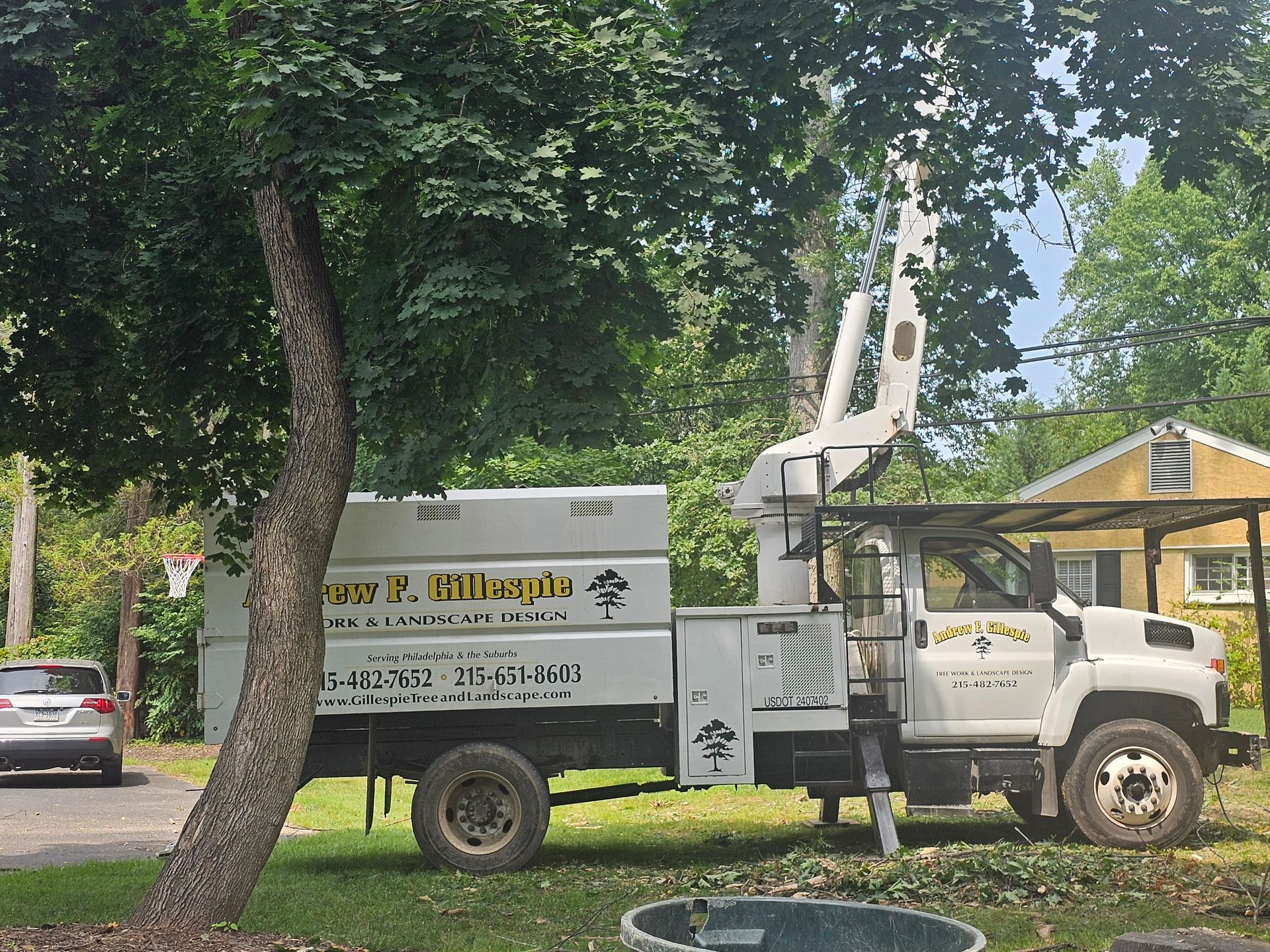 Utility truck with bucket lift parked by a tree in a grassy yard near a house and parked car
