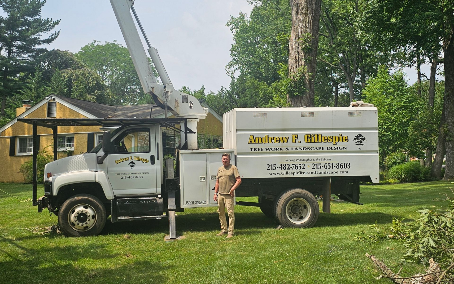 Utility truck parked on grass with raised bucket arm; worker stands beside it near a yellow house.