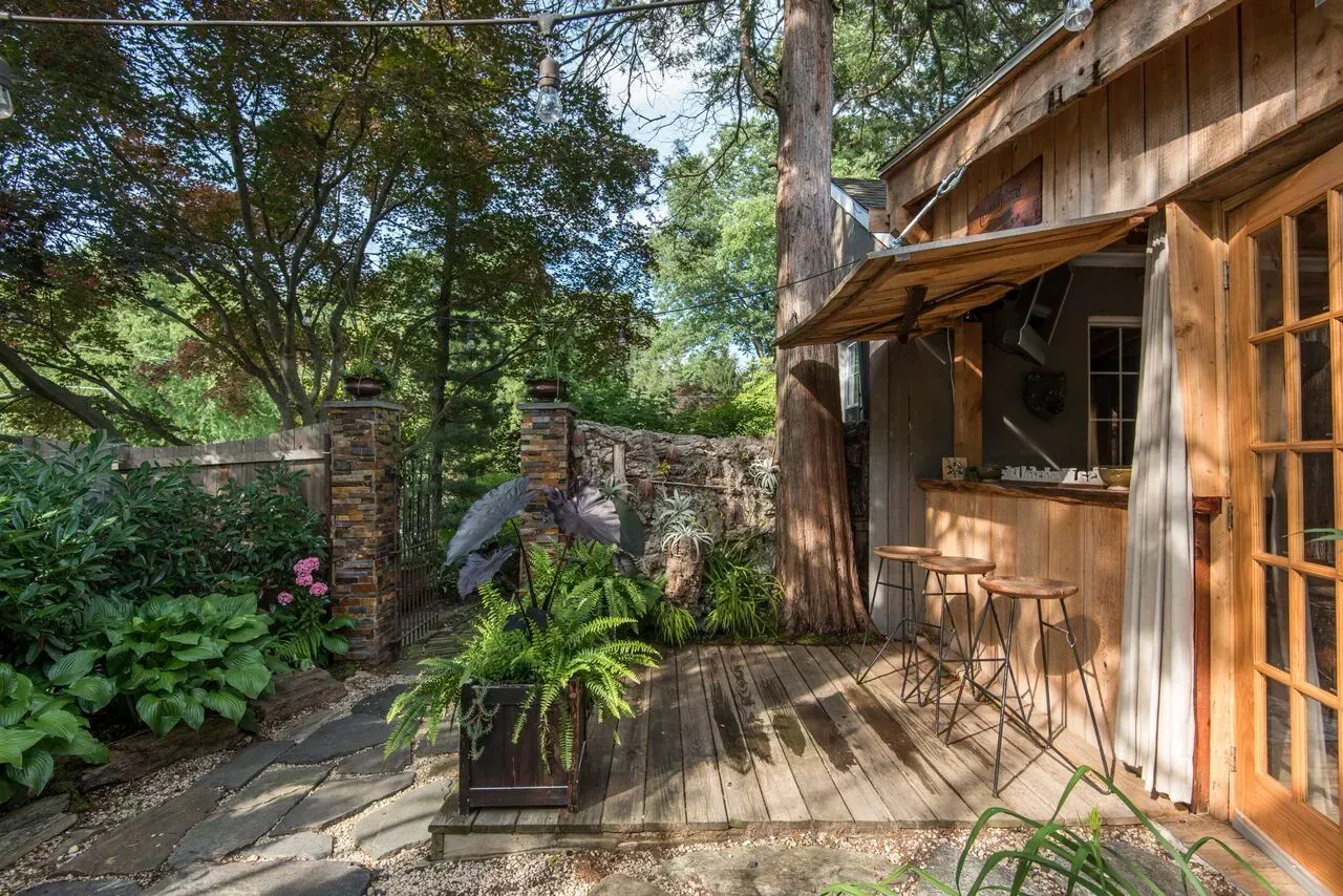 Wooden cabin deck with potted plants, stone wall, and sunlit trees overhead