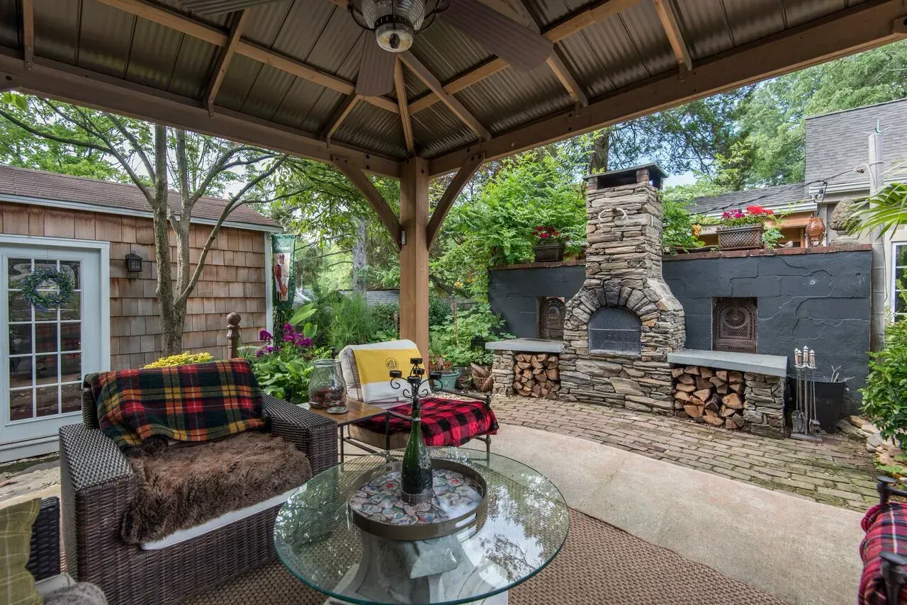 Covered backyard patio with wicker seating, glass table, and stone fireplace against a garden wall