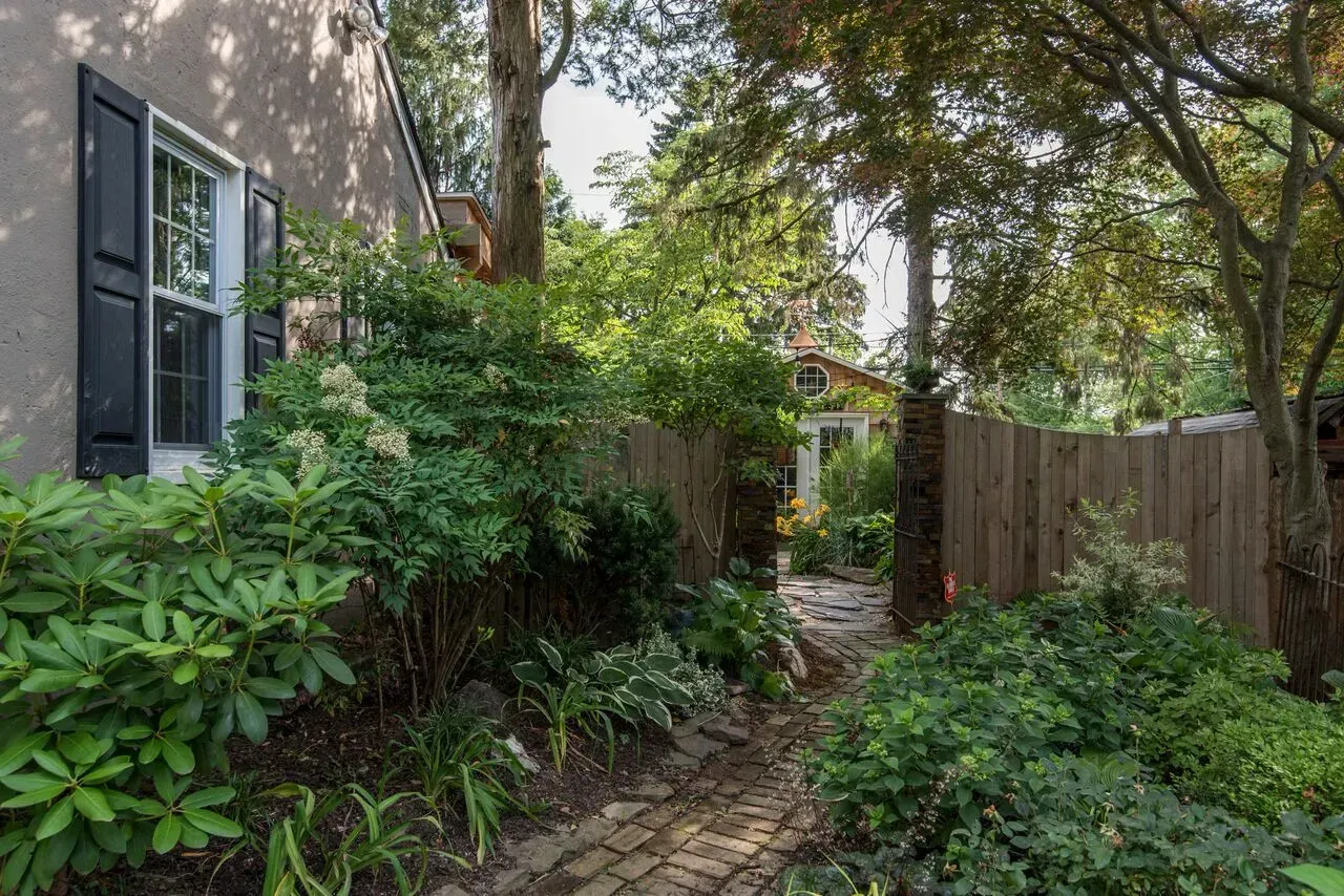 Garden path beside house with lush greenery, wooden fence, and shaded trees