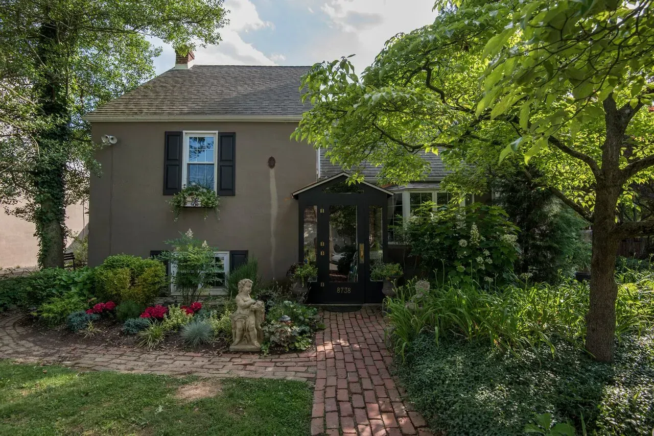 Brick walkway to a tan house with black shutters, front porch, and lush garden trees.