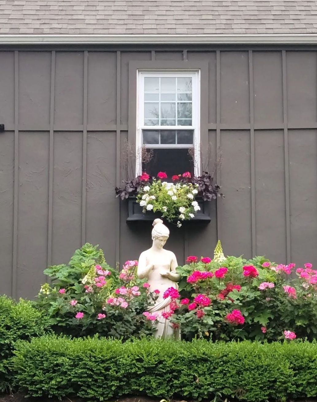 Garden statue amid pink flowers beneath a window on a gray house wall