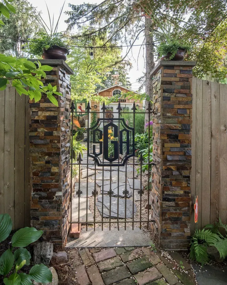 Wrought-iron garden gate between stone pillars, opening to a leafy path and house beyond.