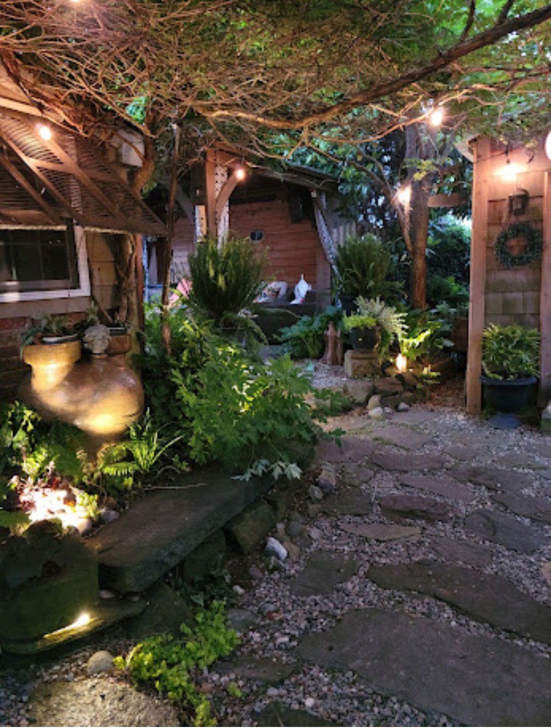 Garden courtyard path with string lights, lush greenery, and rustic stone seating at dusk