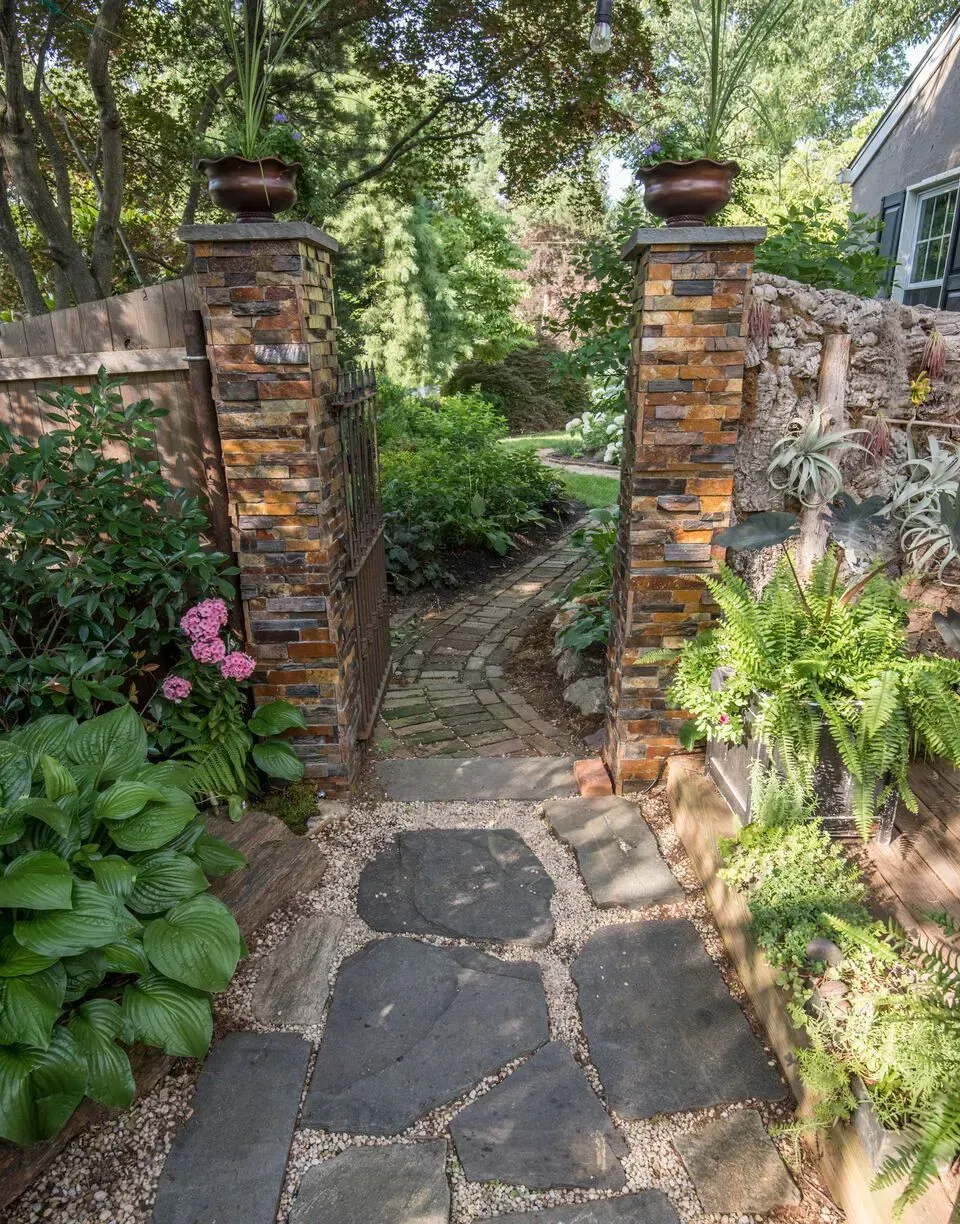 Stone garden path through brick pillars and lush green plants in a shaded backyard entrance