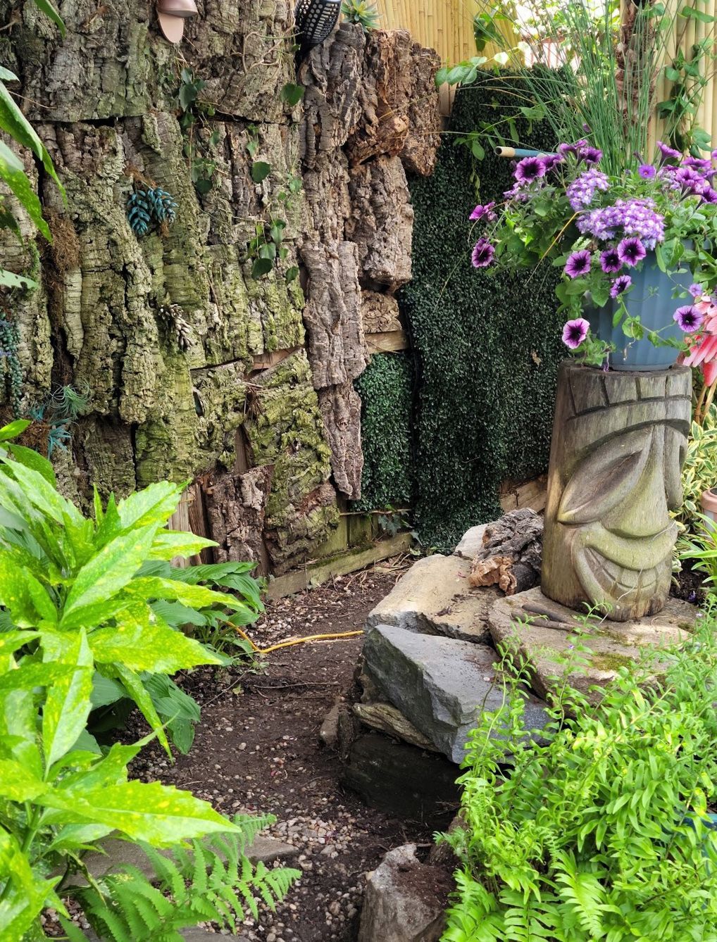 Tropical garden path with mossy rock wall, lush greenery, and a purple flower pot on a stone pedestal