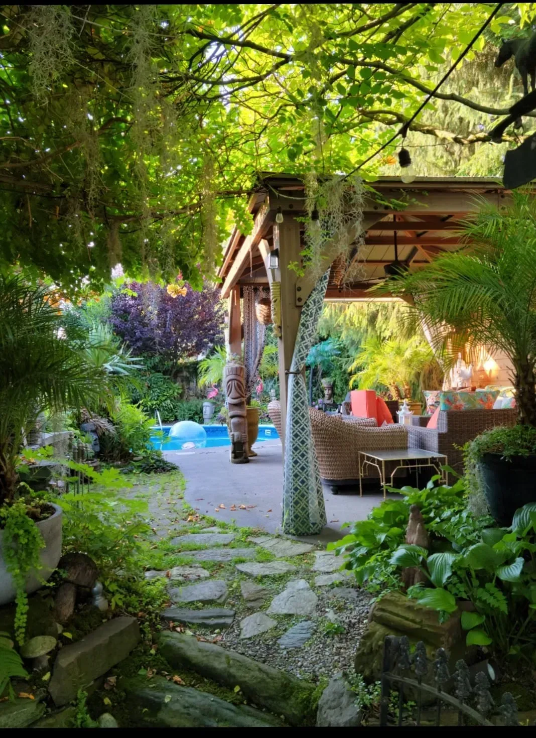 Lush garden patio with stone path, shaded seating, and colorful flowers under a wooden pergola.