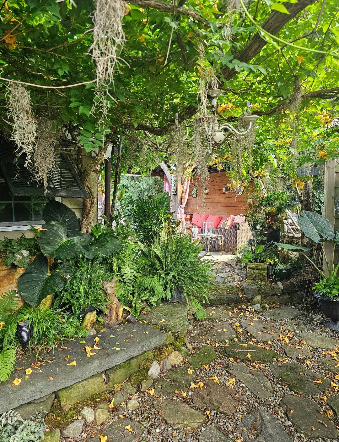 Lush garden path with hanging vines, potted tropical plants, and a stone walkway leading to a red wall