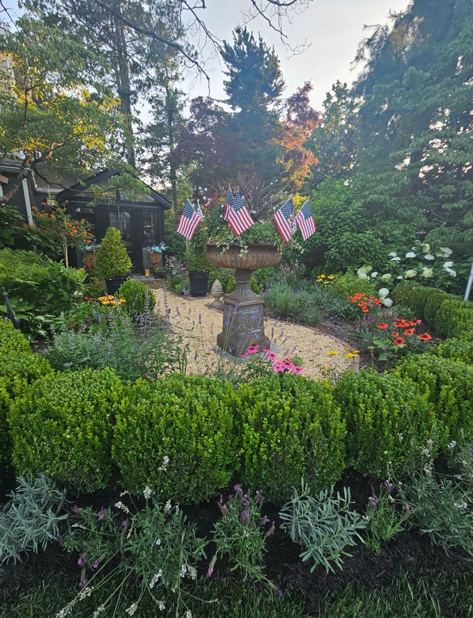 Lush garden fountain with colorful flowers, greenery, and American flags in the background