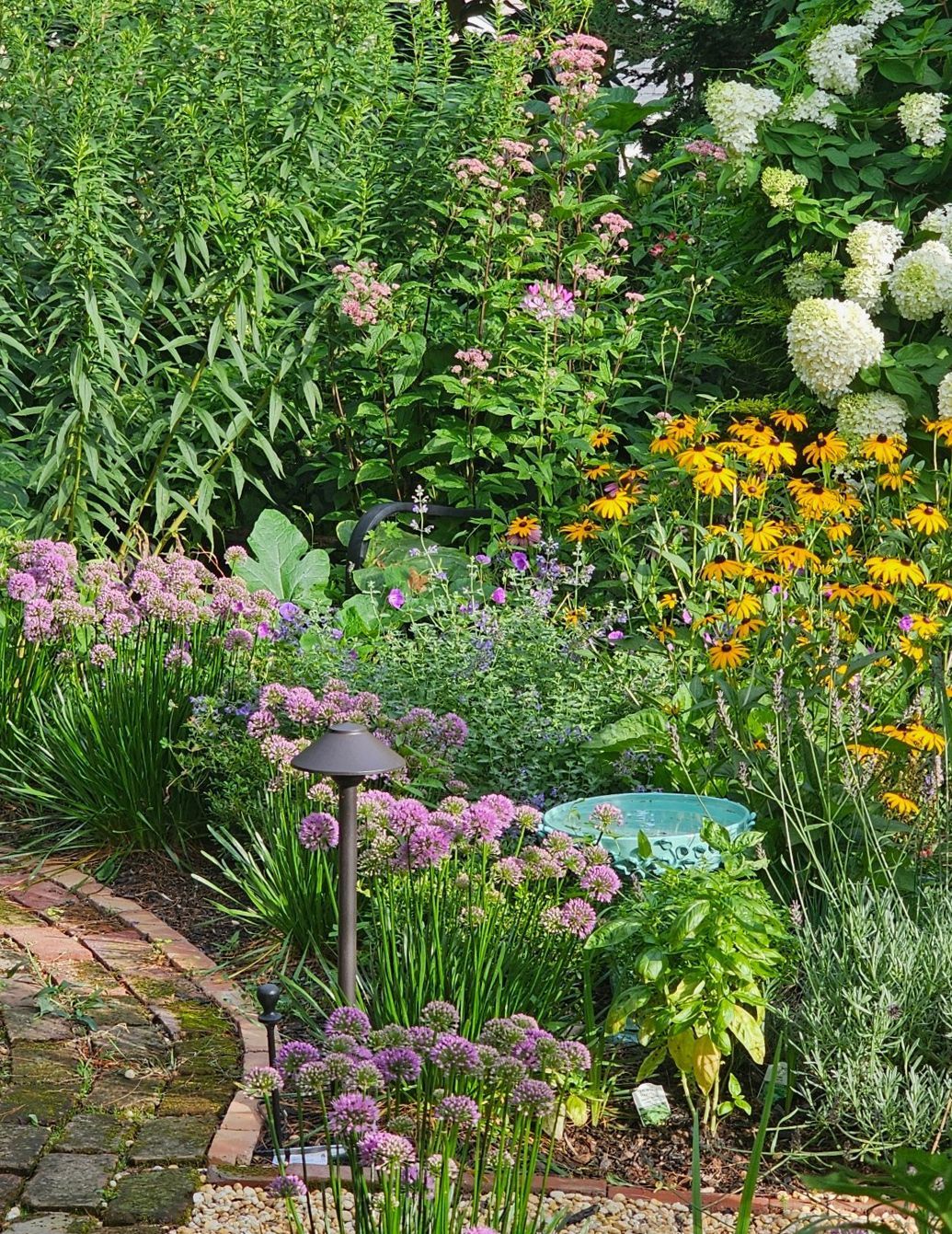 Colorful garden with blooming flowers, lush greenery, stone path, and a small garden lamp