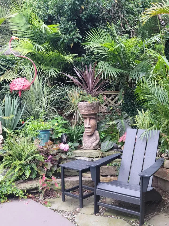 Tropical garden patio with a black chair, small table, and stone face statue among lush plants
