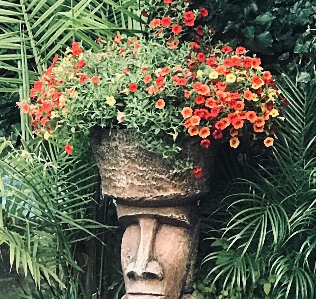 Stone face sculpture topped with red flowers and surrounded by lush green palm fronds