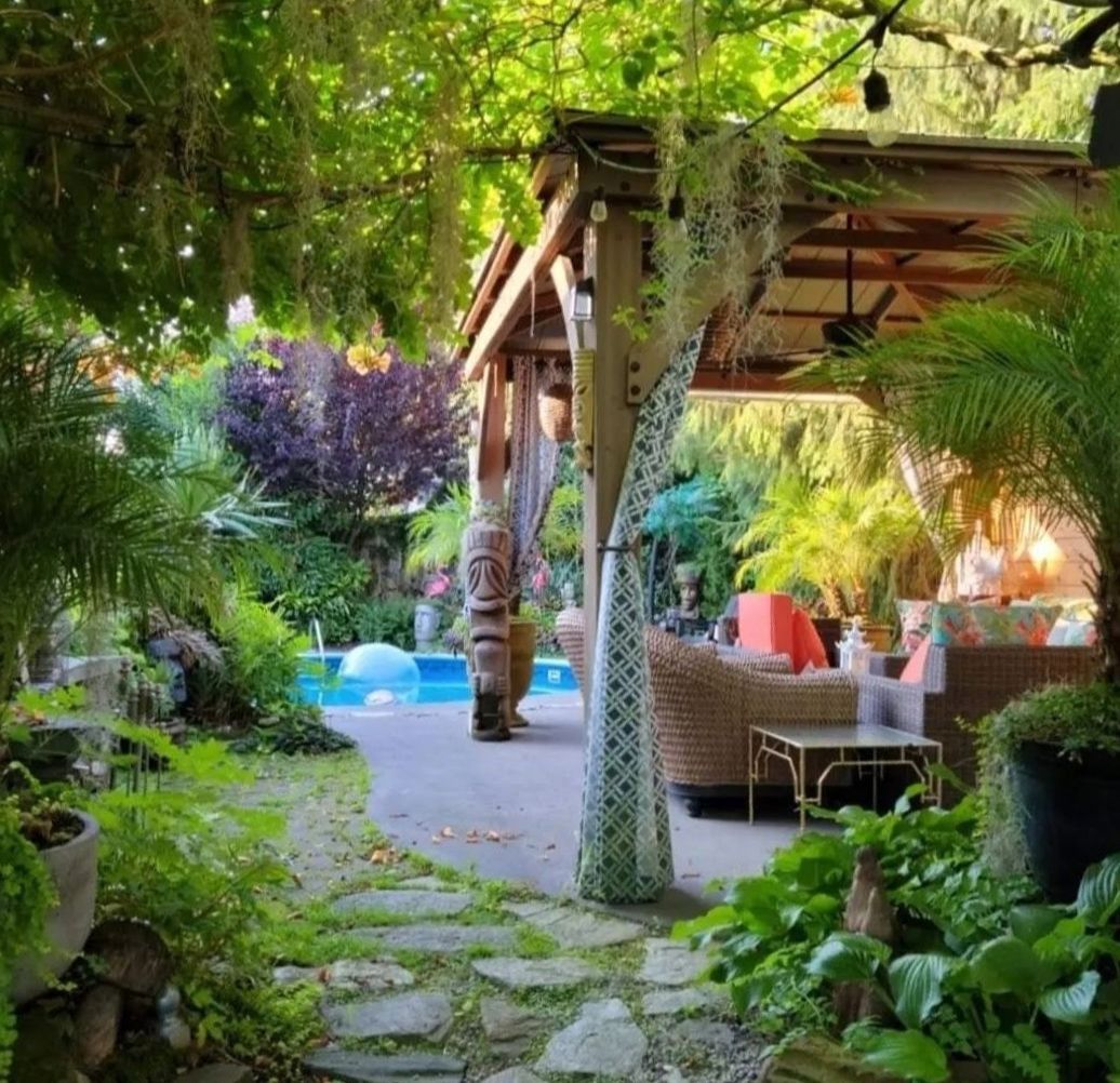 Lush tropical garden patio with stone path, palm trees, and wicker seating under a shaded pergola.
