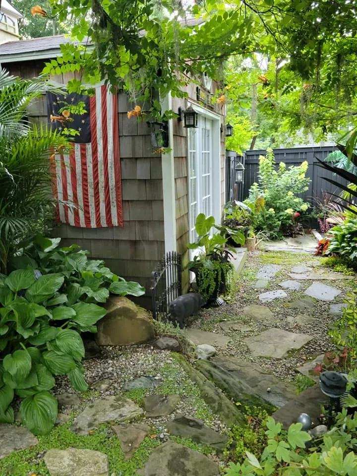Narrow garden path beside a small shed, with lush greenery and a red-striped cloth hanging outside.