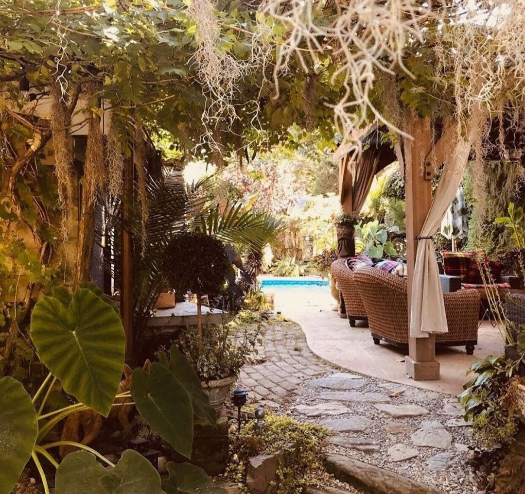 Tropical garden walkway with stone path, hanging vines, wicker chairs, and a bright blue pool beyond