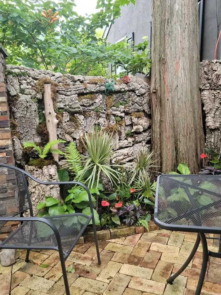 Small stone patio garden with black chairs, brick pavers, plants, and a stone wall beside a tree trunk