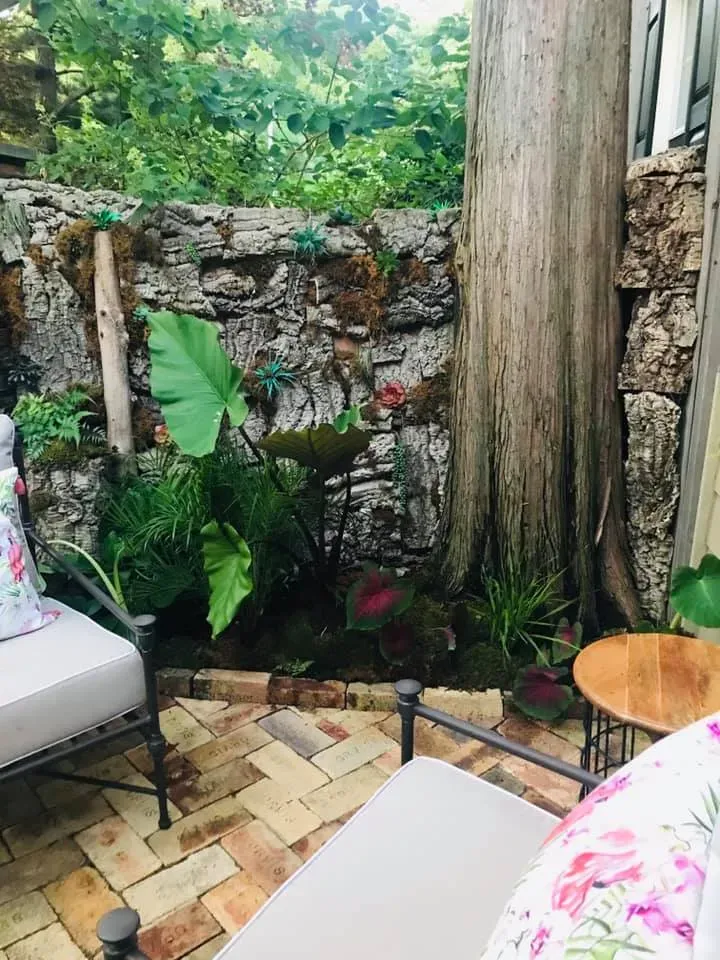 Small outdoor patio with stone wall, tropical plants, tiled floor, and seating with floral cushions
