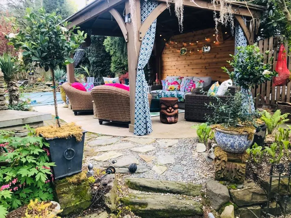 Cozy garden patio with wicker seating, colorful cushions, and a rustic wooden gazebo surrounded by plants