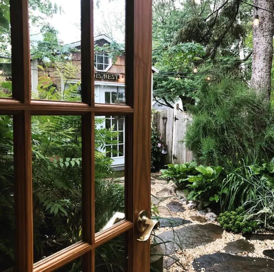 Wooden window overlooking a lush garden path, with a house visible through the trees.