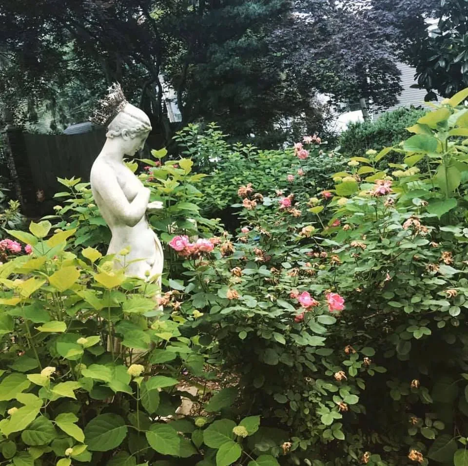 White garden statue among green bushes and pink flowers in a wooded garden