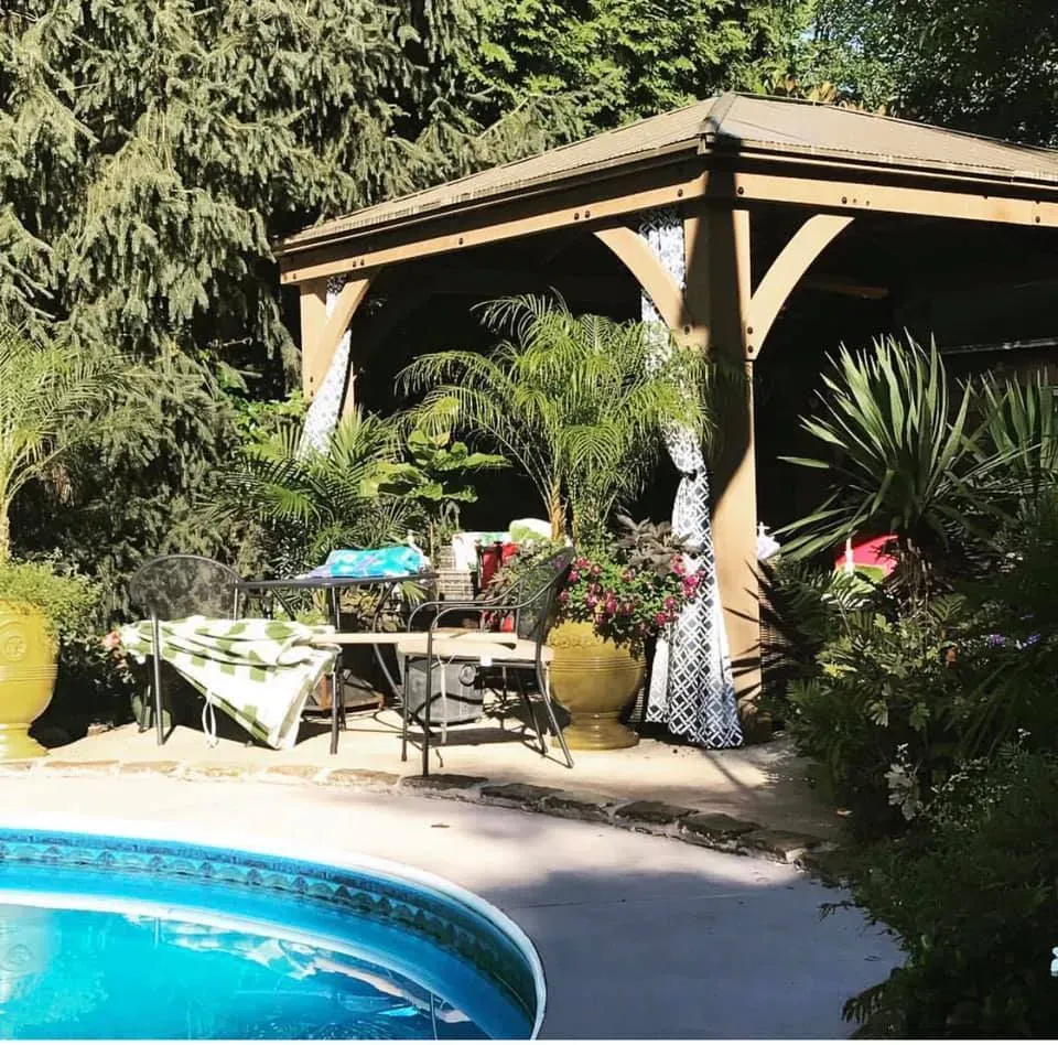 Backyard pool beside a wooden gazebo with potted plants and a table in sunlight