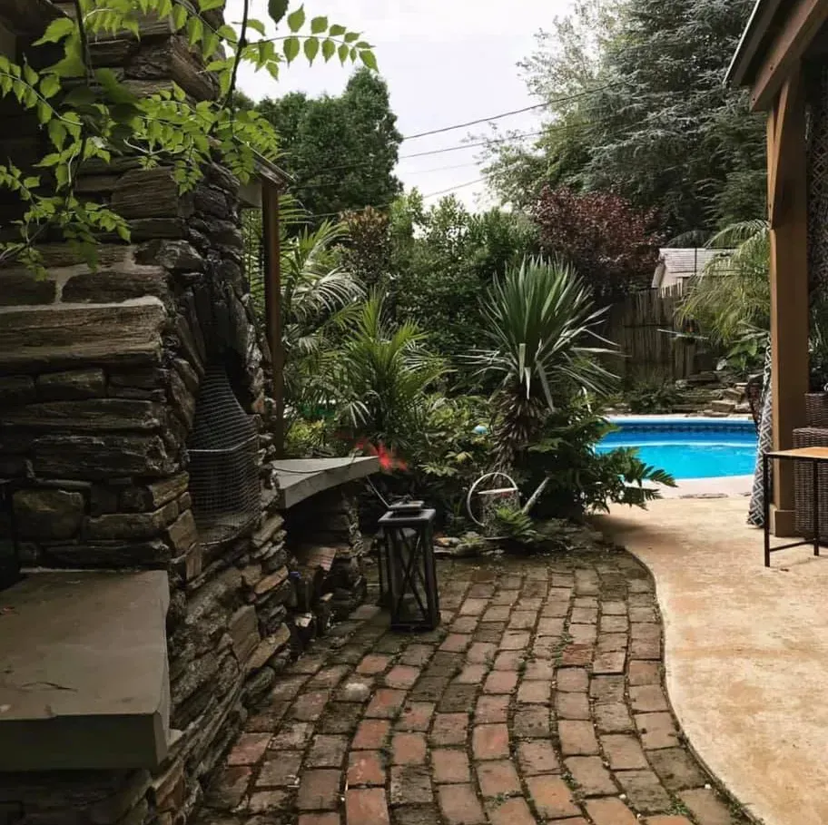 Brick path leading to a blue backyard pool beside lush greenery and a stone wall.