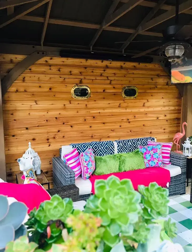 Cozy patio lounge with wicker sofas, bright pink and green cushions, and wooden wall décor under a roof.
