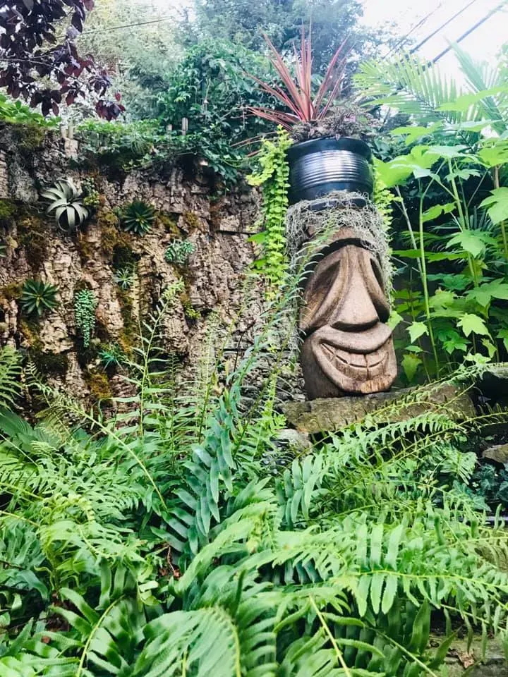 Tropical garden with lush ferns, a carved wooden face, and a black pot planter on a stone wall