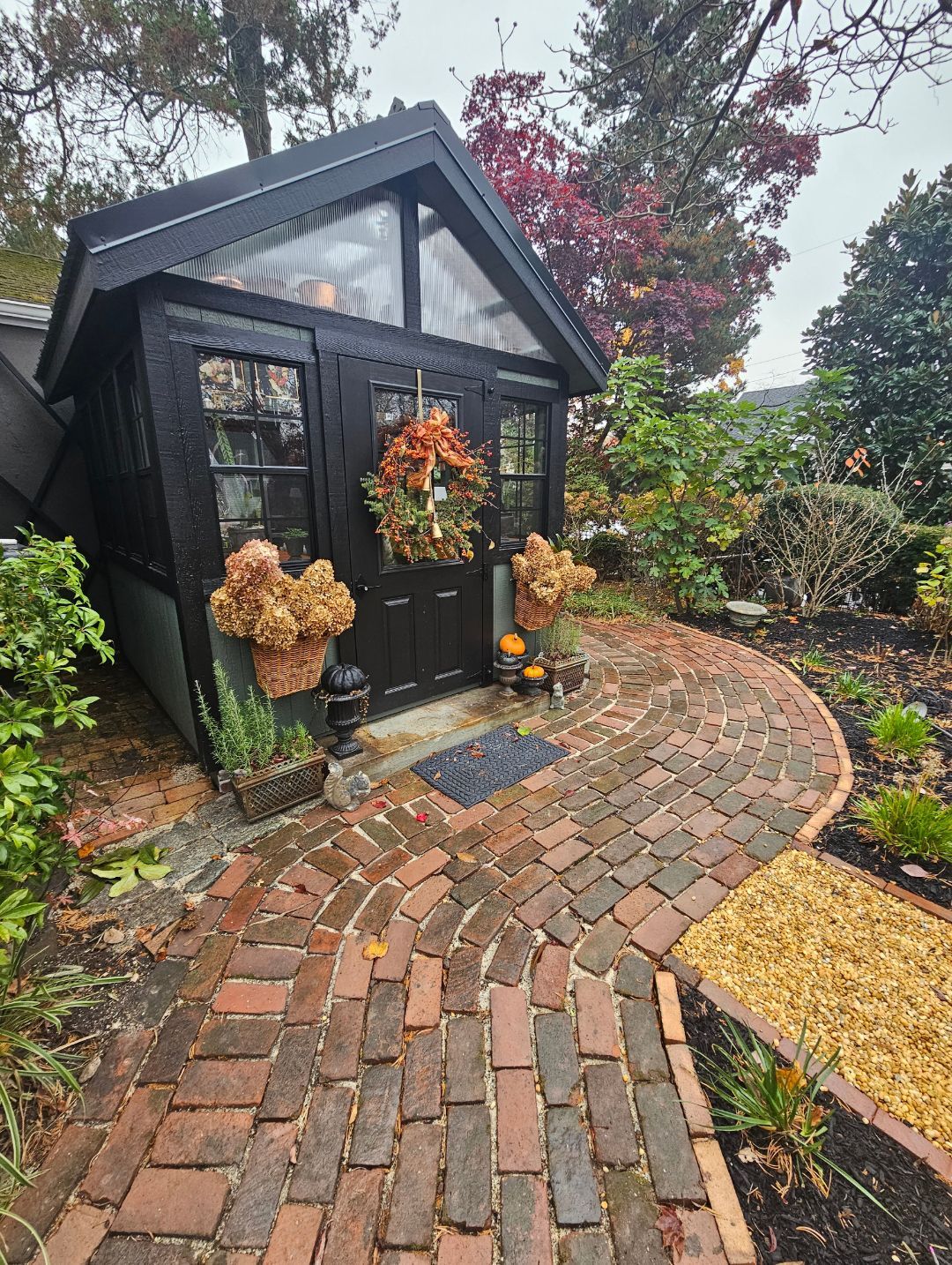 Black garden shed with a wreath beside a curving brick path in an autumn yard