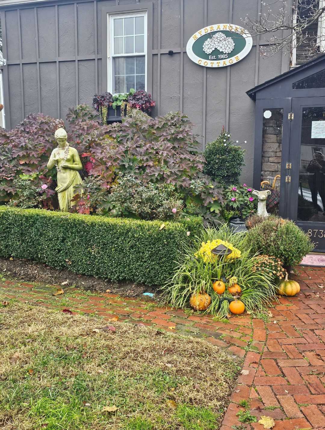 Garden display with a statue, hedges, pumpkins, and a brick path beside a gray building