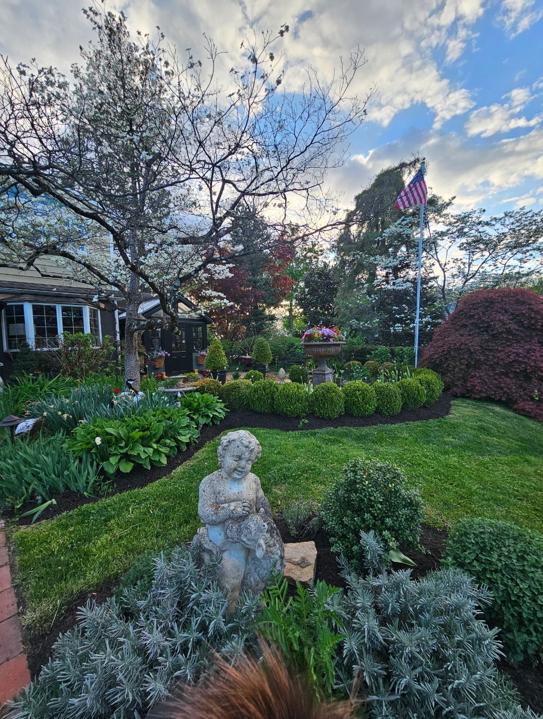 Stone statue in a landscaped garden with shrubs, trees, and a cloudy evening sky