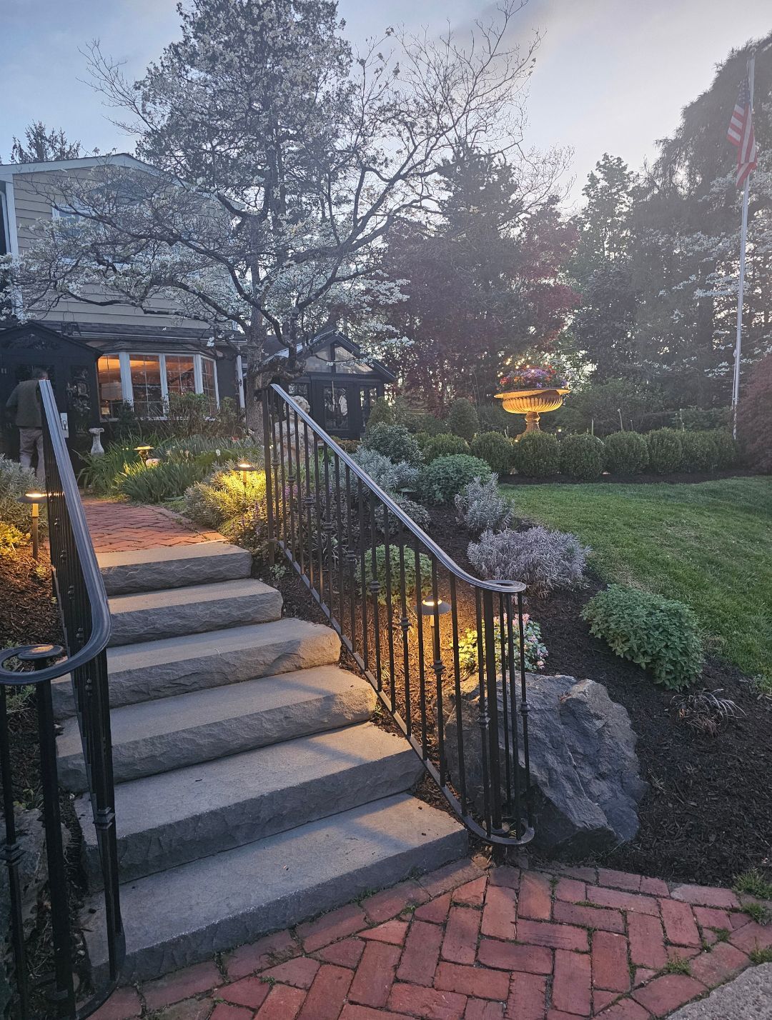 Stone steps with black railings lead to a lit house garden at dusk, with red brick patio and greenery.