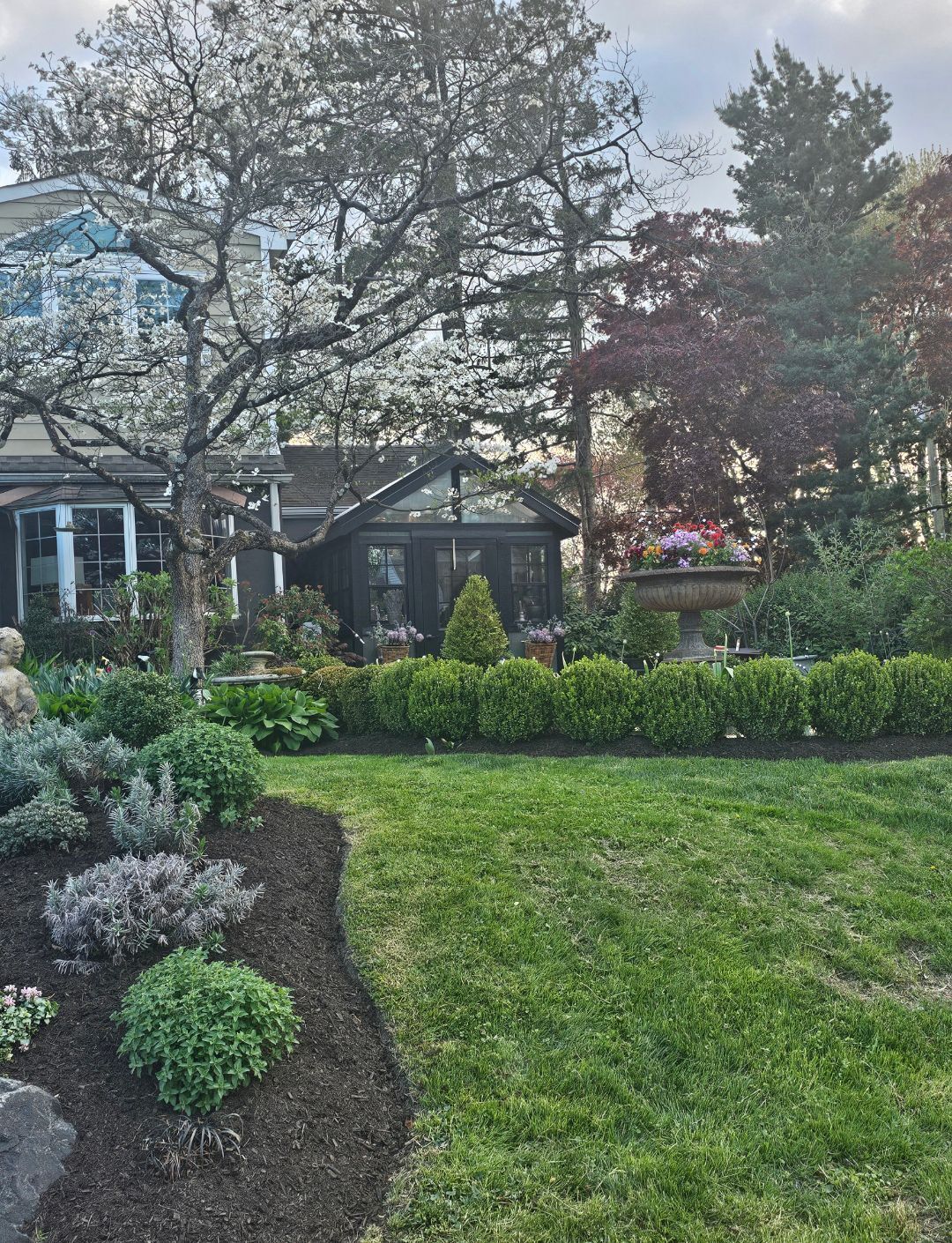 Landscaped garden with green lawn, shrubs, and trees beside a house under an overcast sky