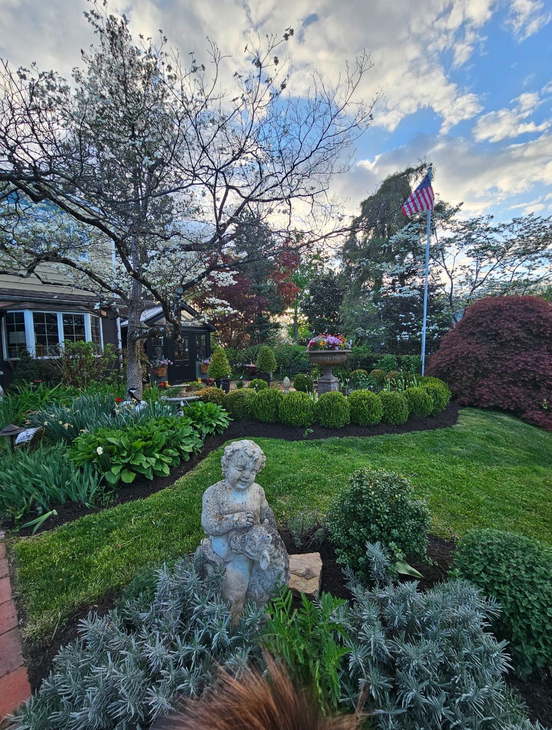 Flower-filled front garden with a stone child statue, manicured shrubs, and an American flag at sunset