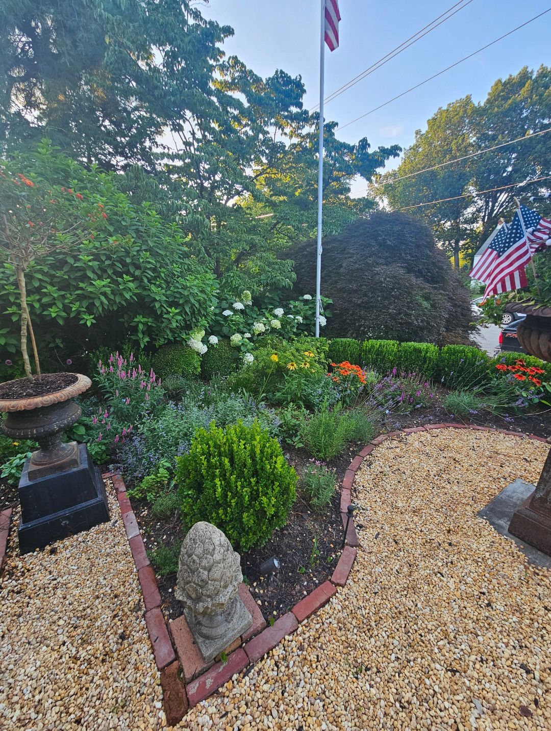 Front garden with flowers, gravel path, flagpole, and brick edging under trees